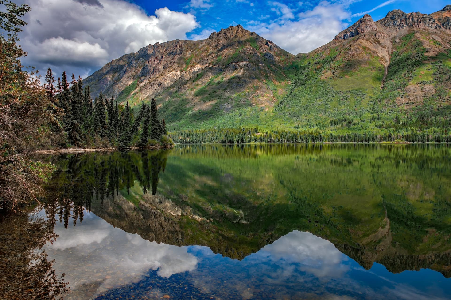 Green mountain slopes and jagged peaks reflected in a still Yukon lake under blue sky and white clouds